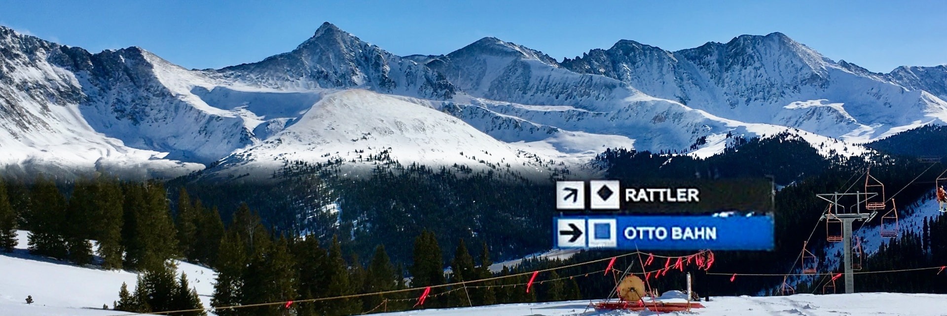 Snow-covered Colorado peaks above a ski run marked by Rattler black diamond and Otto Bahn blue square trail signage